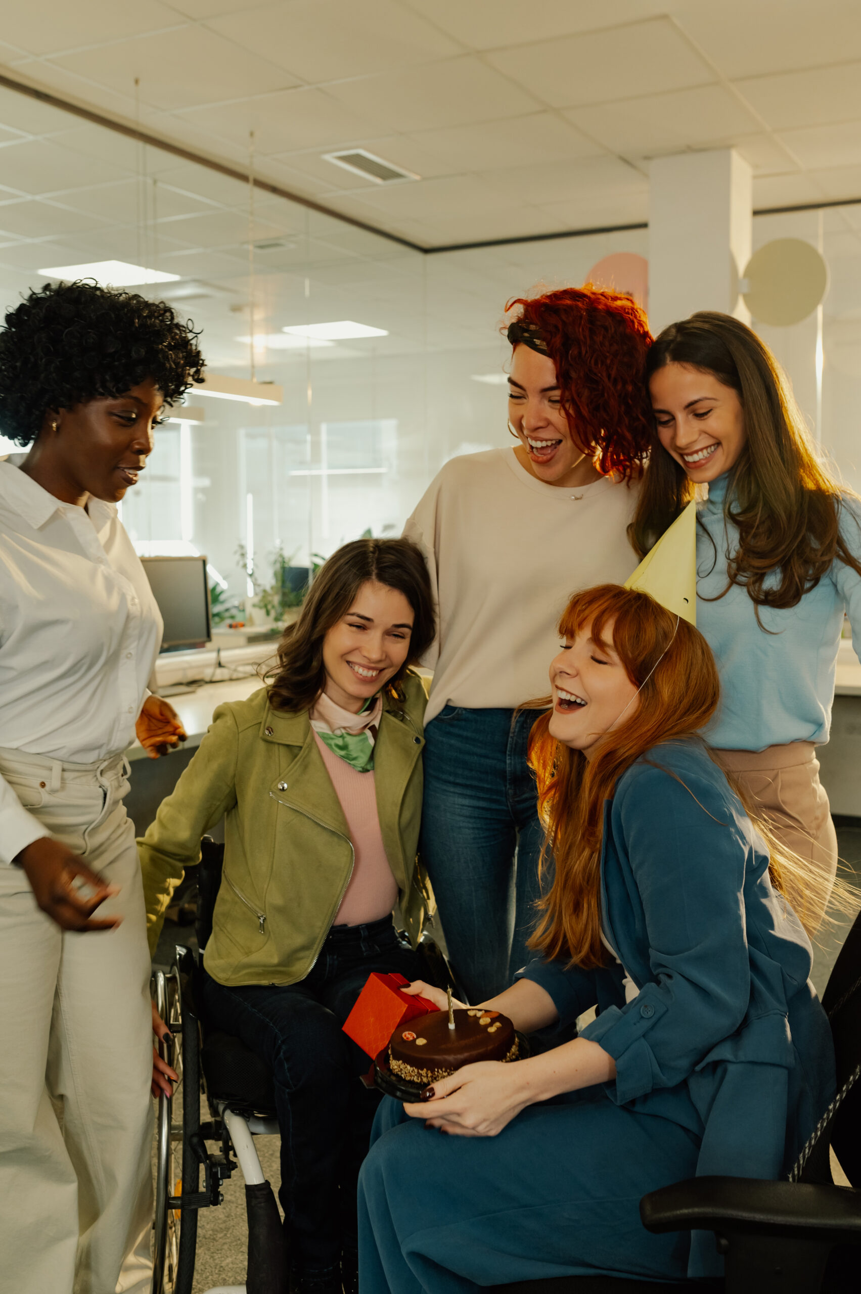 A happy young female diverse female colleagues having a birthday surprise party with a businesswoman sitting at the office with a cake. A cheerful businesswoman celebrating birthday at workspace.